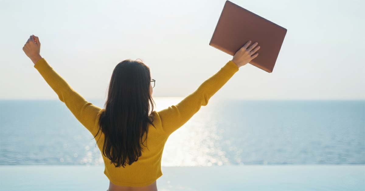 Rear view of a woman in a yellow ribbed top raising her arms in a celebratory pose while holding a laptop, overlooking an infinity pool and the sunlit ocean.