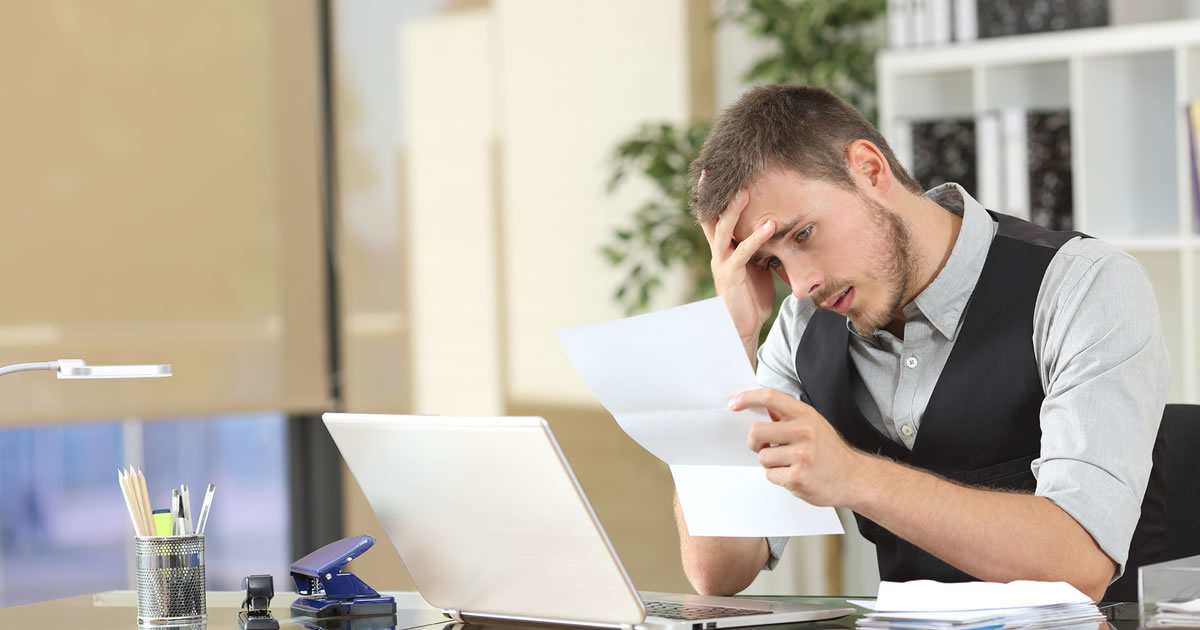 Businessman at desk with his hand to his head and serious expression as he looks at piece of paper