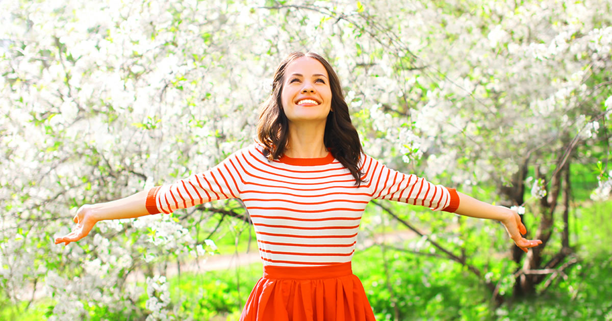 Smiling woman standing outdoors in front of trees with her arms outstretched