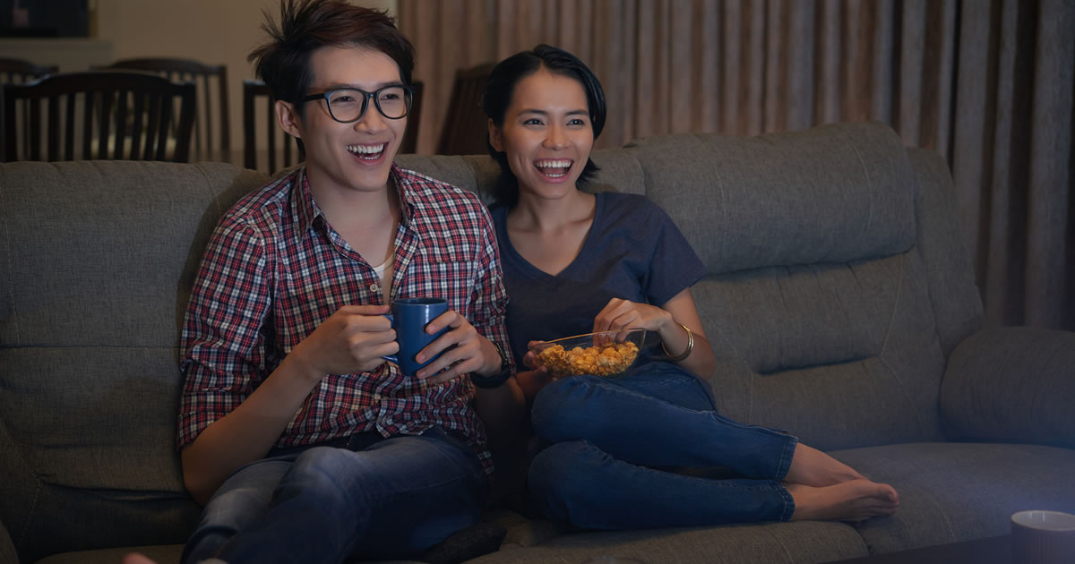 Duas pessoas sorridentes com bebida e lanche sentadas juntas no sofá assistindo à televisão