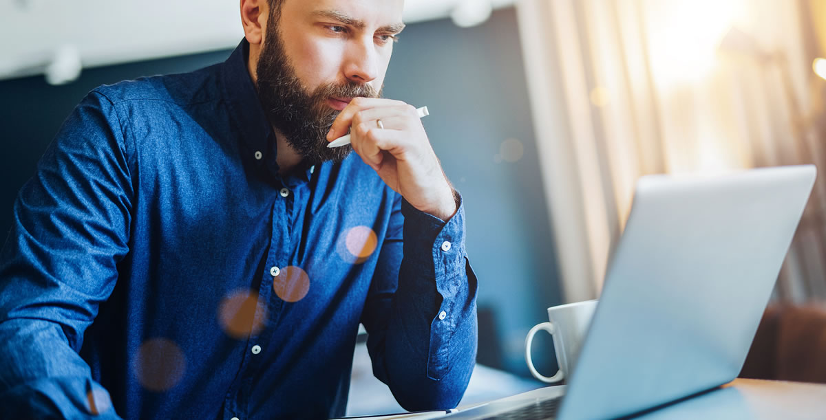 Man sitting at desk with thoughtful expression while holding pen and looking at laptop screen