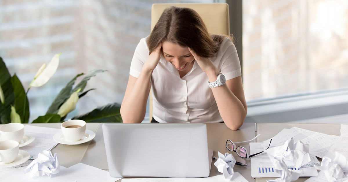 Woman with her head in her hands sitting at a desk surrounded by crumpled up papers