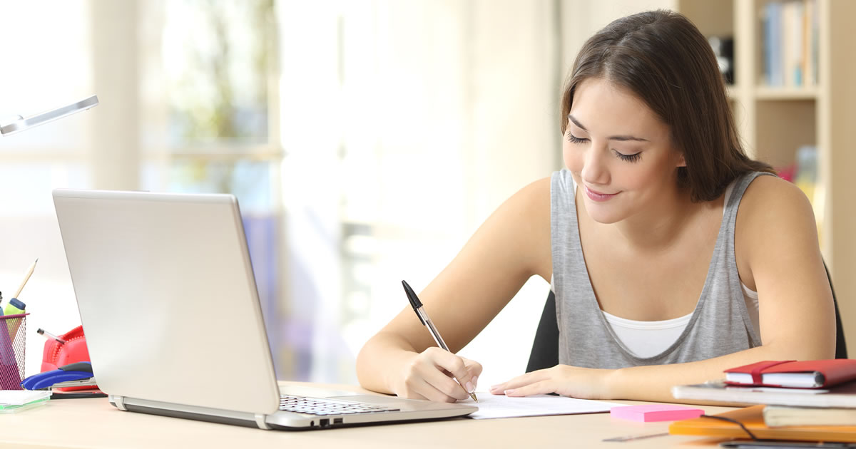 Woman smiling to herself as she writes a note while sitting at a desk in front of a laptop