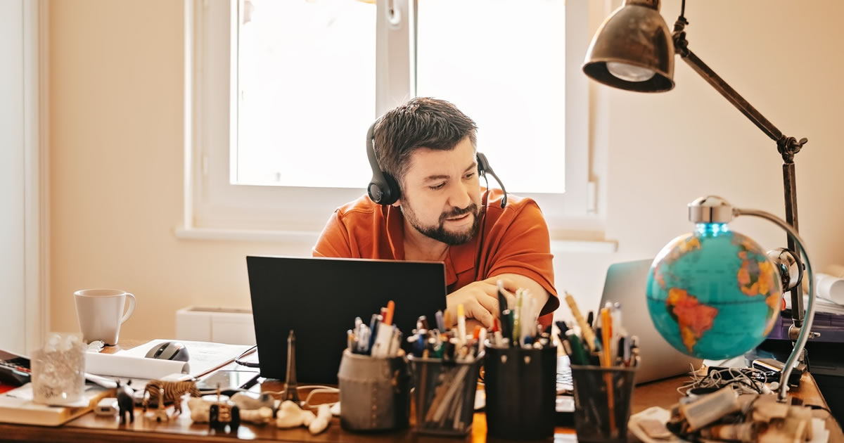 Male writer with headphones and microphone having conference call online sitting at home office with two laptops