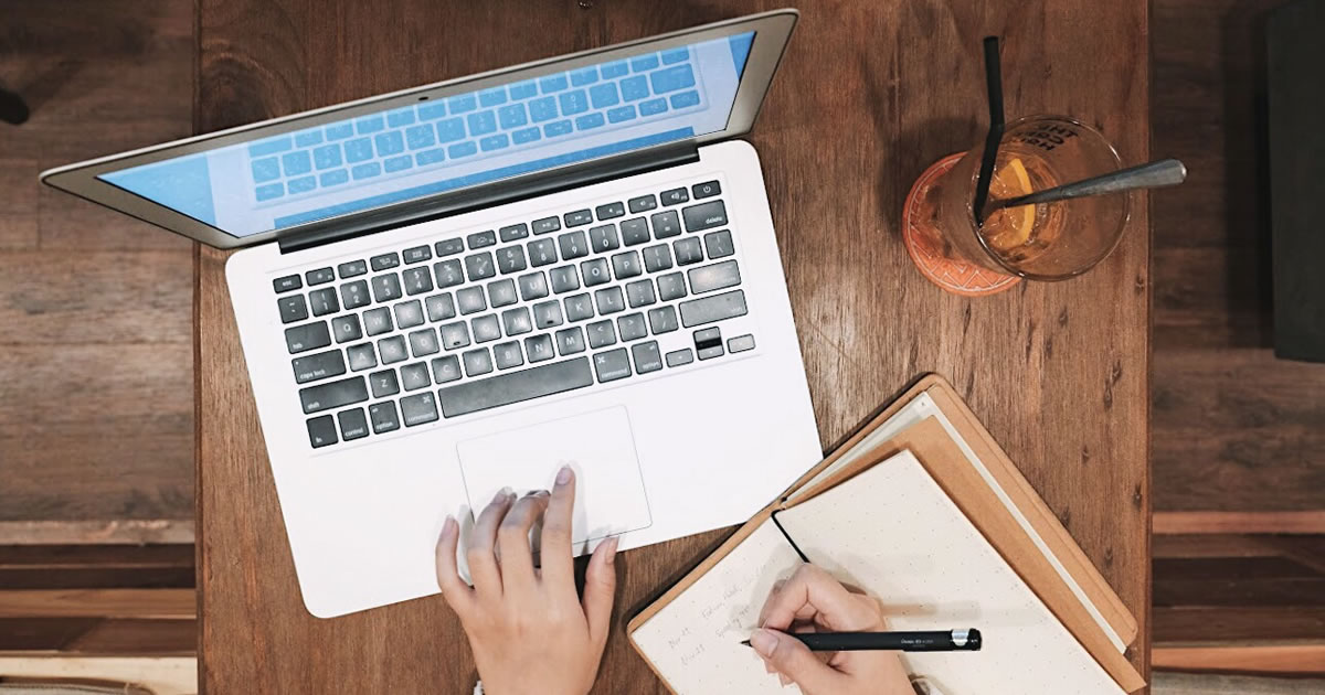 Overhead view of computer laptop with a writer adding notes into a notebook