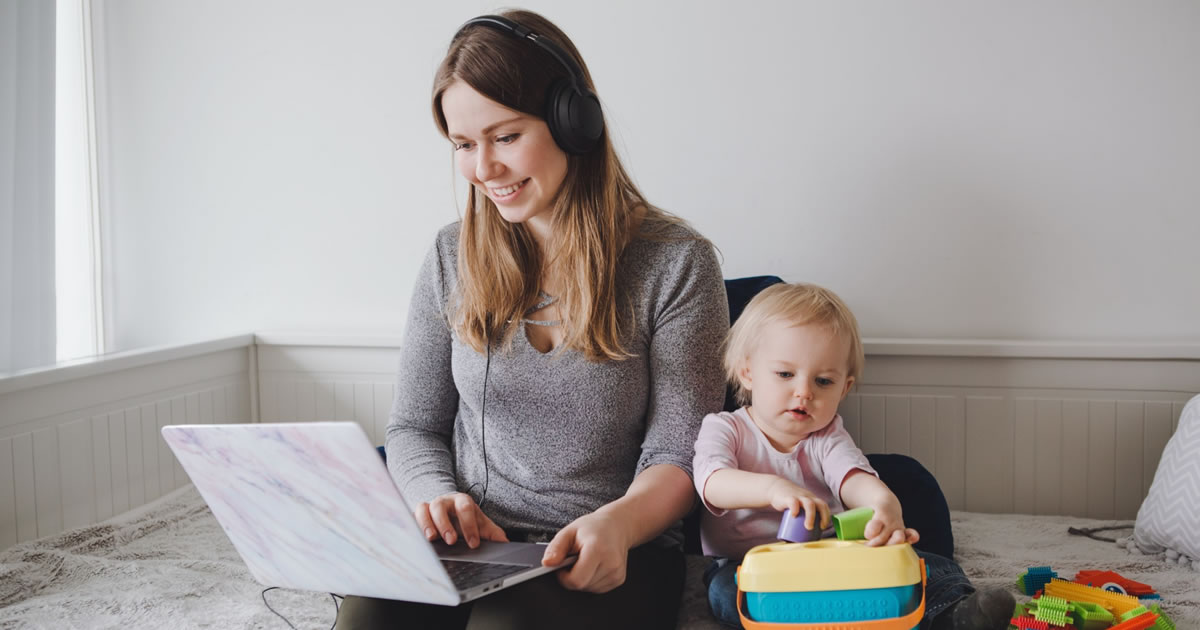 Smiling woman wearing headphones and using laptop while sitting next to young child holding a toy