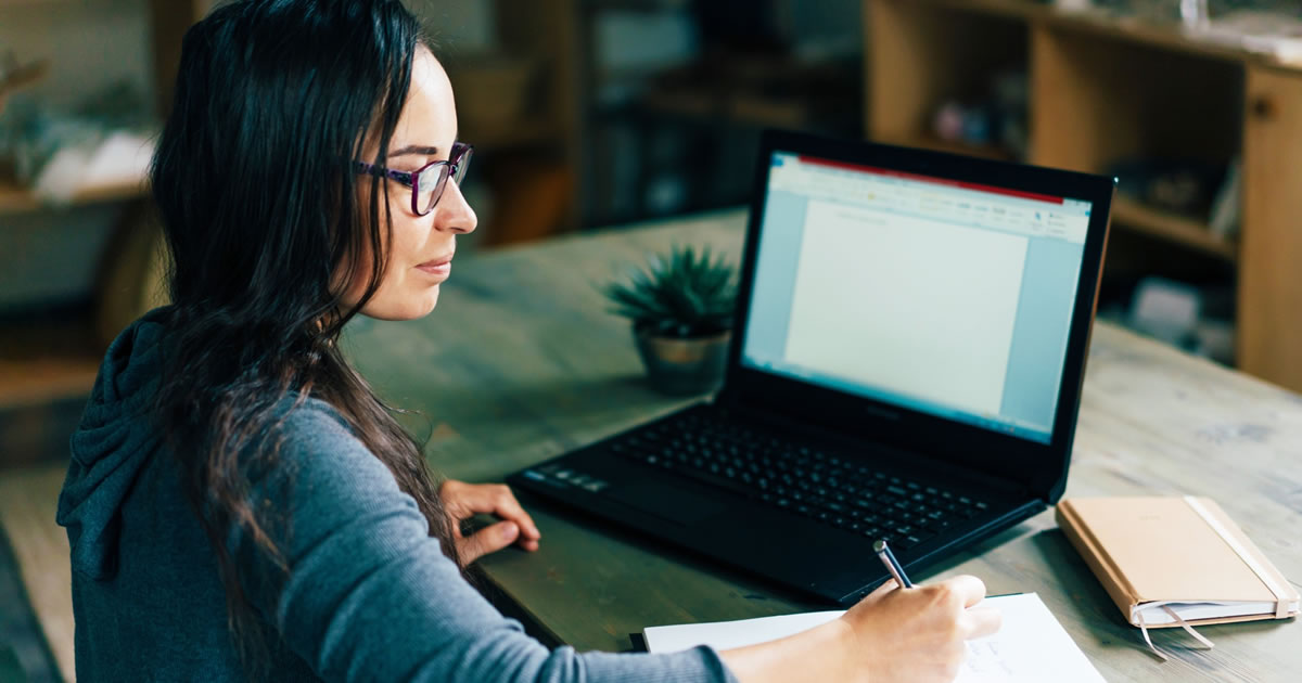 Woman at home working at desk with laptop, pen, and notepad