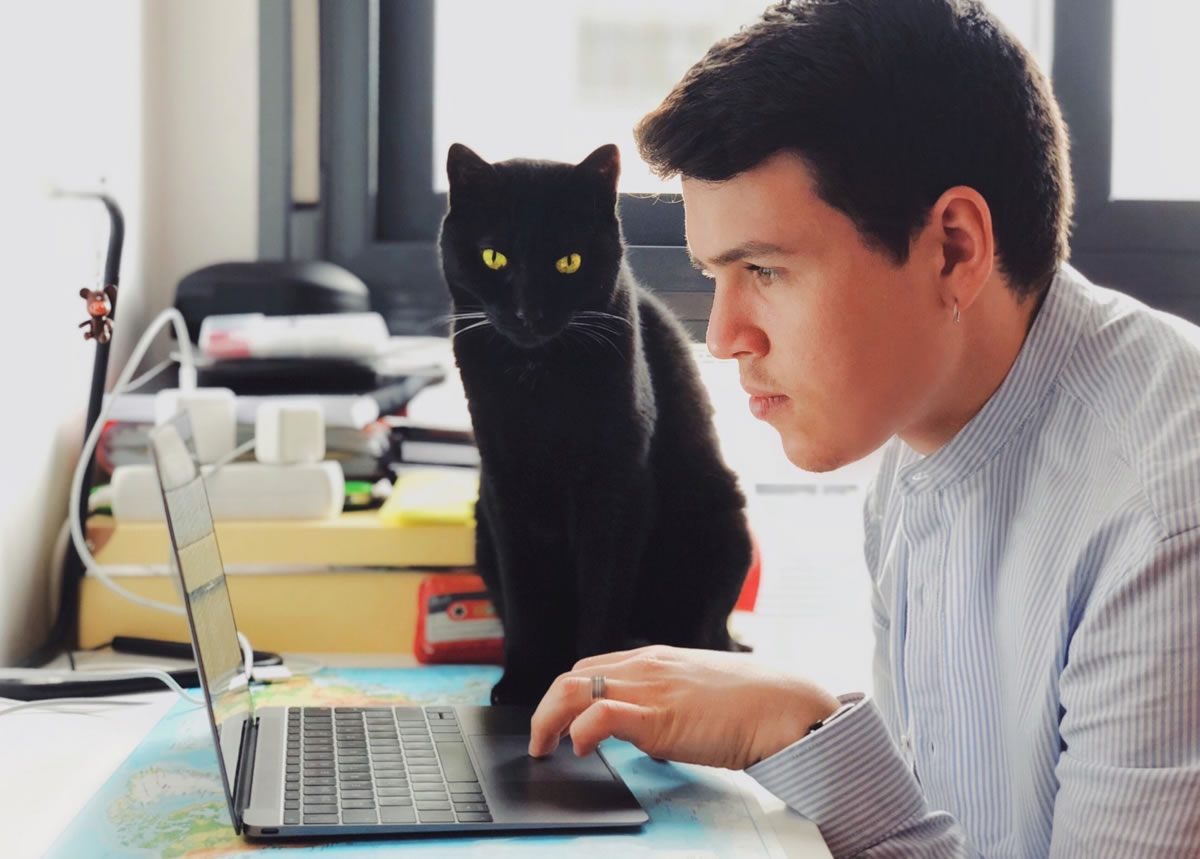 Man working on laptop with black cat sitting on desk beside it