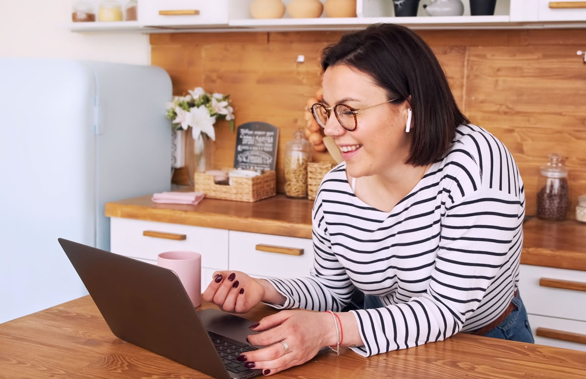 Smiling woman wearing wireless earphones and looking at laptop while leaning on kitchen counter next to coffee mug