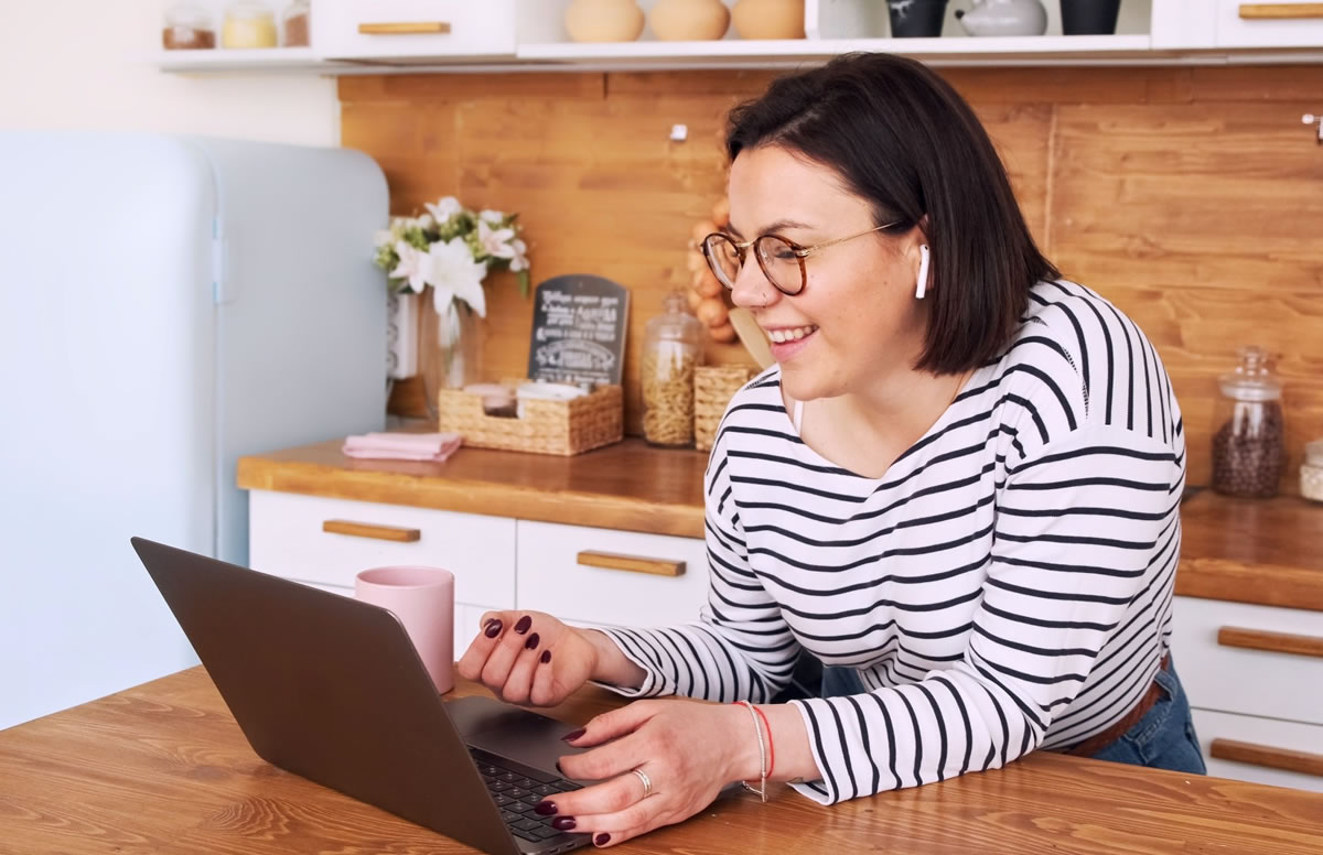 Mulher sorridente usando fones de ouvido sem fio e olhando para o laptop enquanto se apoia no balcão da cozinha ao lado de uma caneca de café