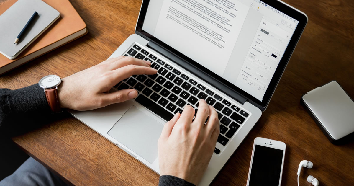 Overhead closeup photo of a man's hands on a wooden desk typing on a laptop next to a phone, headphones, and notebooks