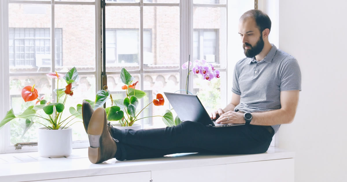 Homem reclinado confortavelmente no assento da janela, escrevendo no laptop