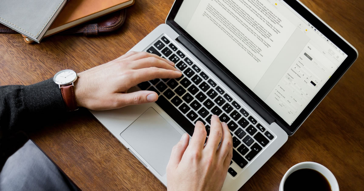 Man sitting at a wooden desk in home office writing on a laptop computer with a coffee mug