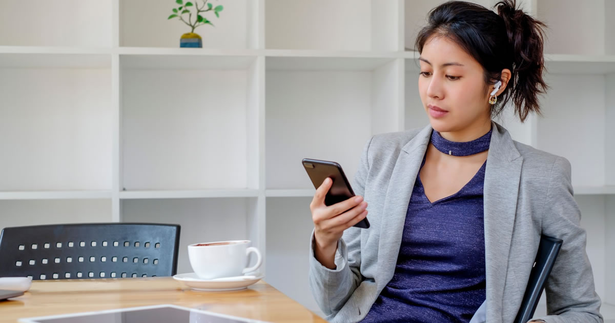 Businesswoman with coffee sitting at office table looking at smartphone while wearing wireless earphones