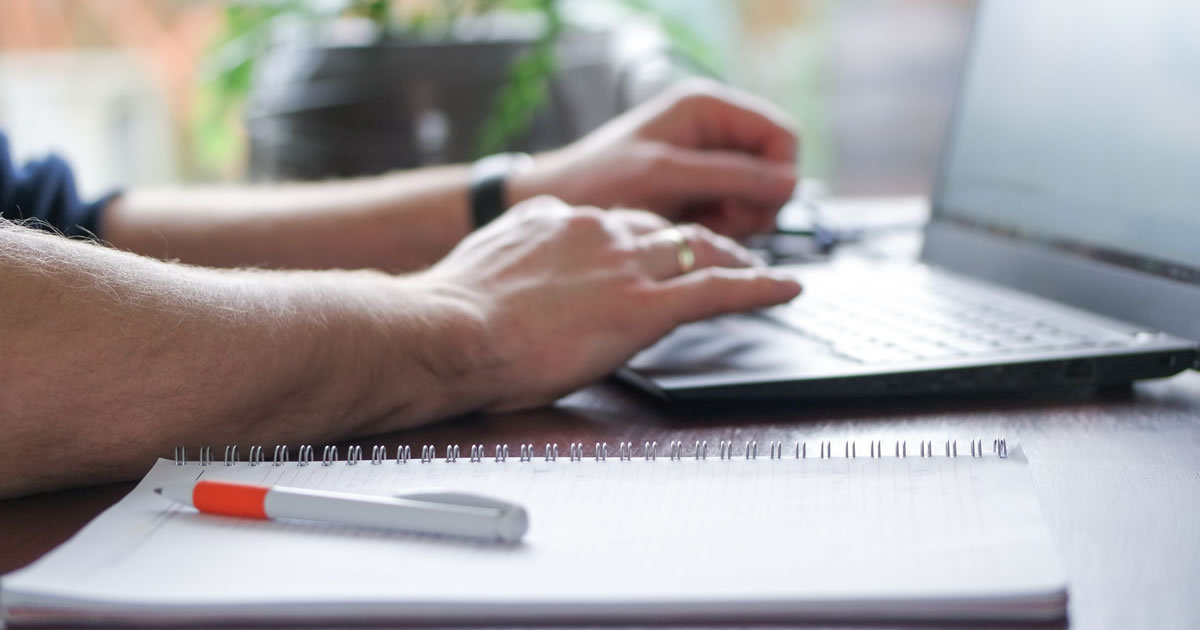 man writing on laptop with planning notebook nearby