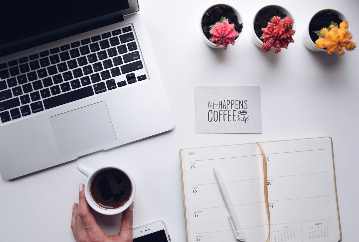 Overhead shot of a desk with a laptop computer, mobile phone, weekly planner, coffee cup, and a card saying LIFE HAPPENS COFFEE HELPS