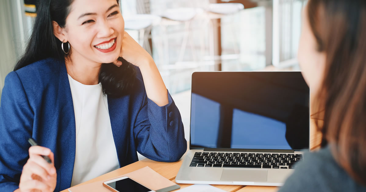 Two smiling businesswomen sitting at desk talking with laptop in front of them