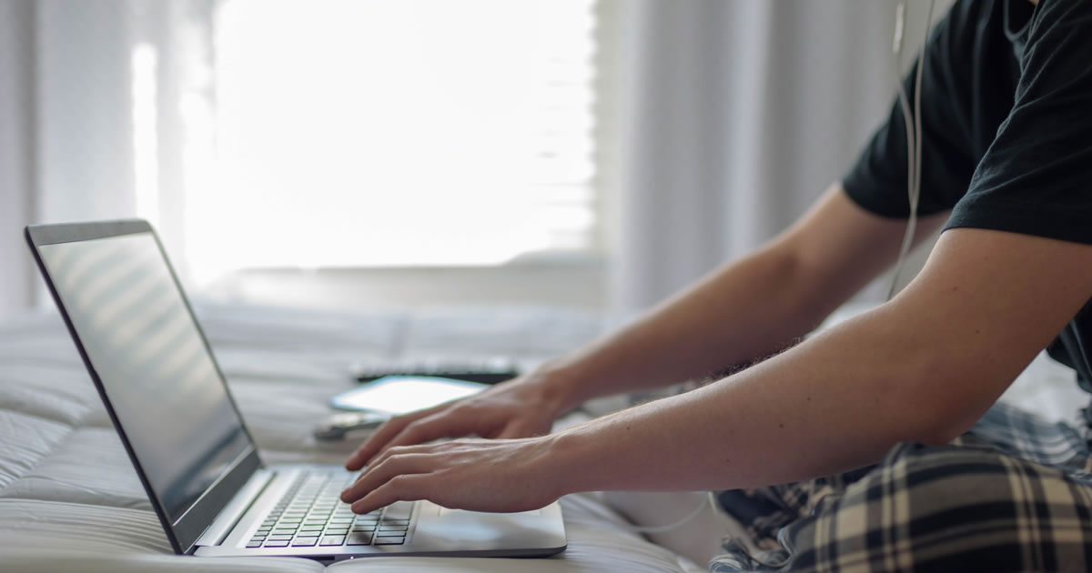 Young man wearing earbuds sitting on bed working on laptop