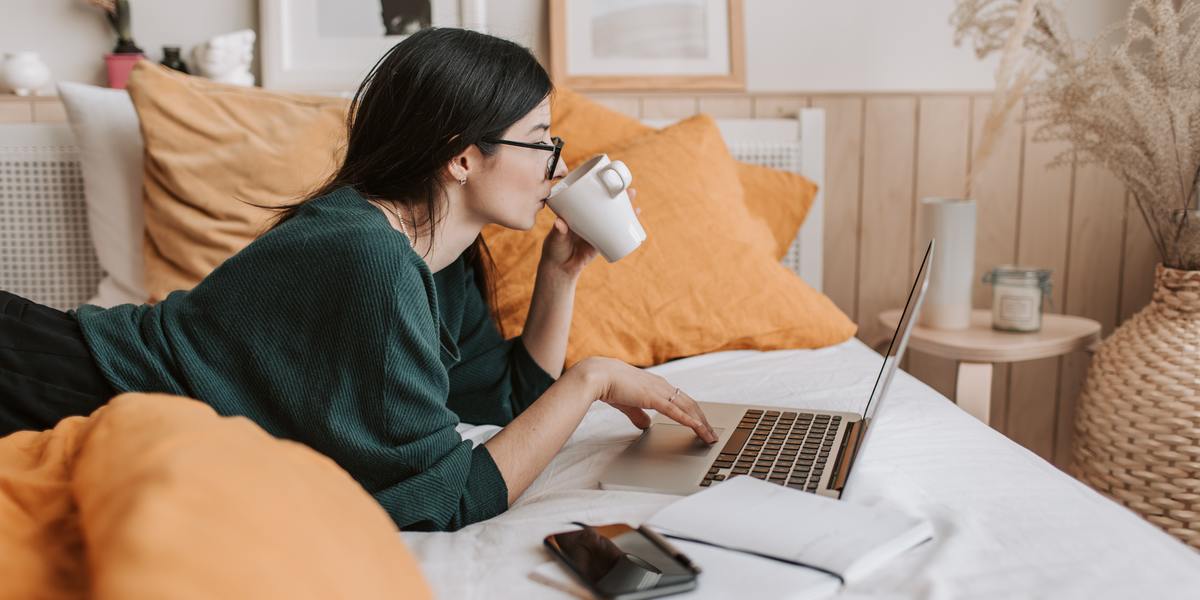 Young woman drinking coffee and writing on laptop laying in bed