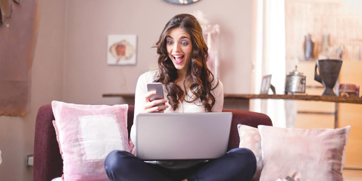 Young woman happy and surprised looking at her phone and writing on laptop