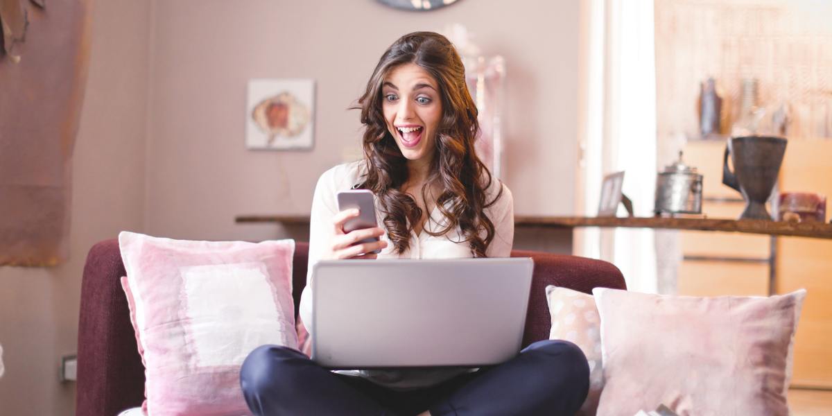 Mulher jovem, feliz e surpresa, olhando para o celular e escrevendo no laptop