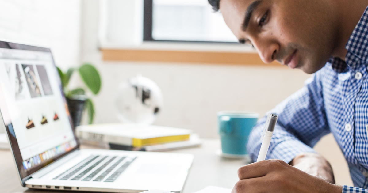 Homem escrevendo em um caderno na frente de um laptop sobre a mesa