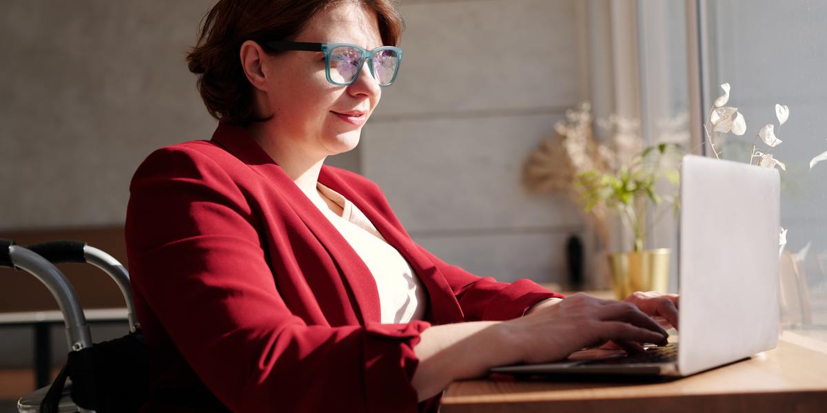 A senhora de blazer vermelho, usando óculos, escrevendo em seu laptop