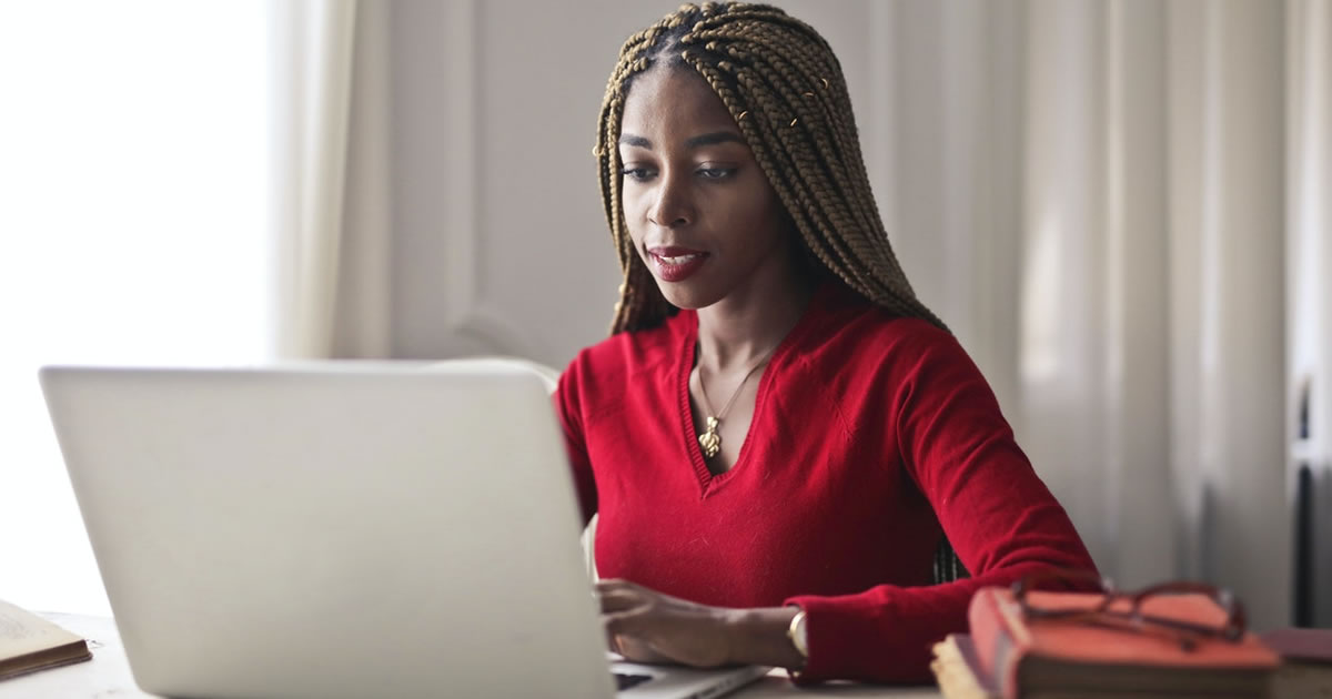 Mulher de blusa vermelha escrevendo em seu laptop sentada em uma mesa