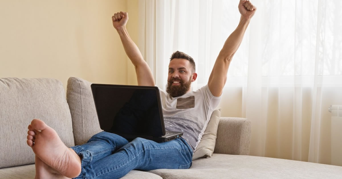 Smiling man on couch looking at laptop with arms raised in air