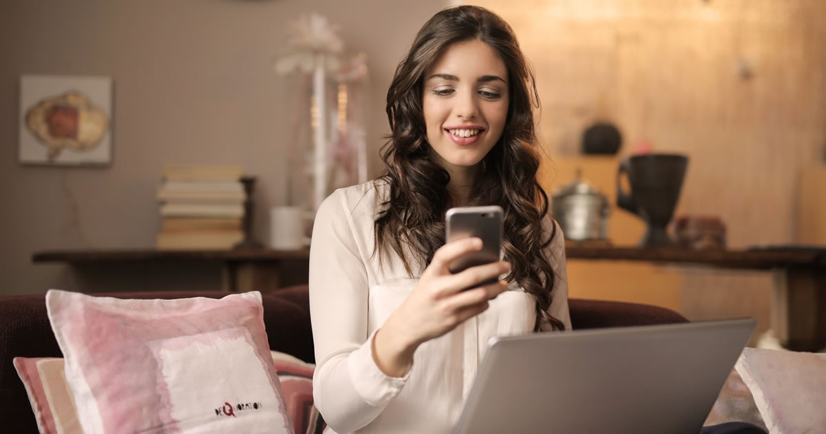 Woman sitting on sofa studying her phone with a laptop on her lap