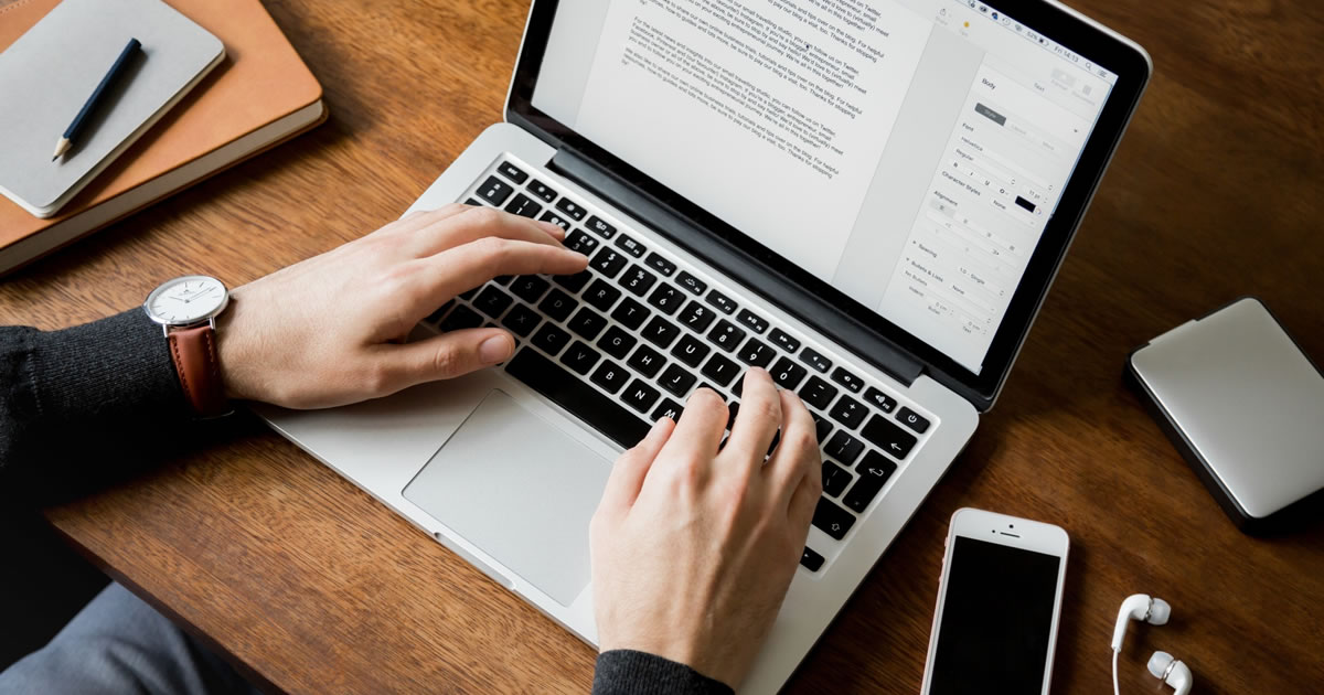 Closeup of a man's hands at a wooden desk typing on a laptop computer with a phone, headphones, notebooks, pencil, and hard drive on the desk