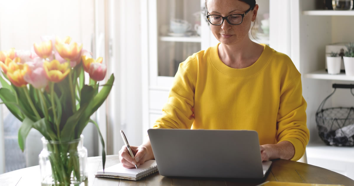 Woman in yellow sweater sitting at a table using laptop computer and writing notes in notebook