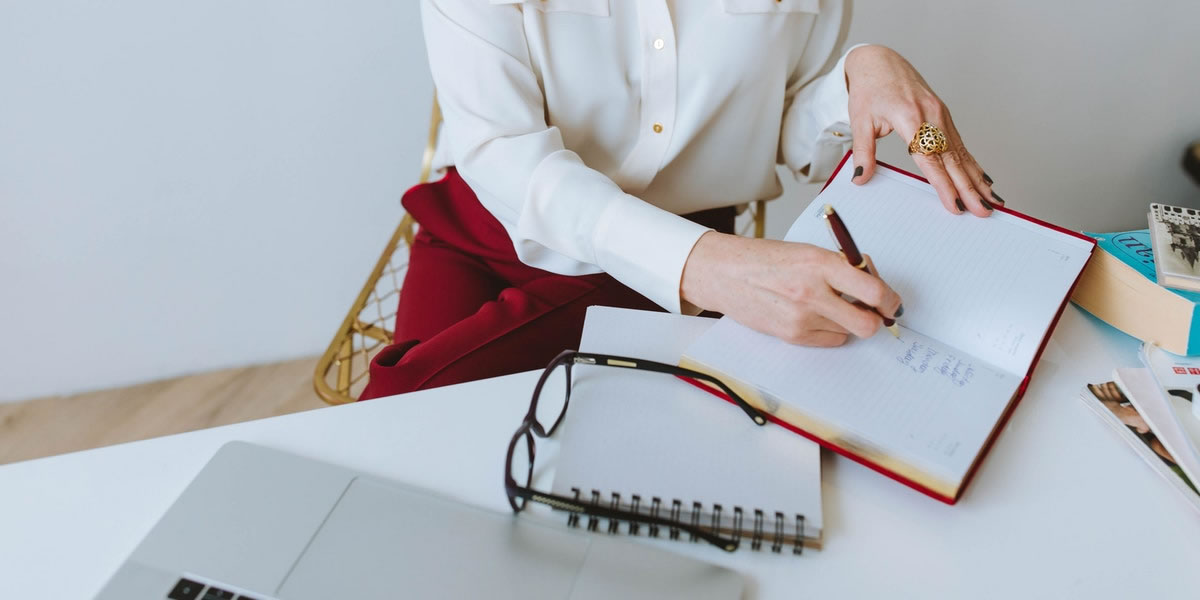 A woman's hands holding a pen at a desk with a notebook, eyeglasses, and a laptop computer