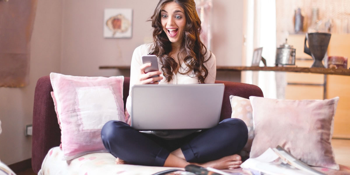 Mulher feliz olhando para o celular enquanto está sentada no sofá com um laptop