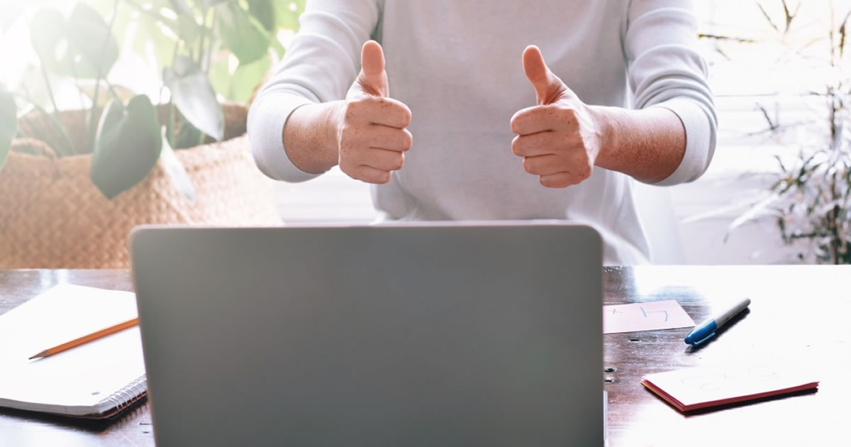 Closeup of person sitting at table with open laptop, notepad, and pen while giving two thumbs up