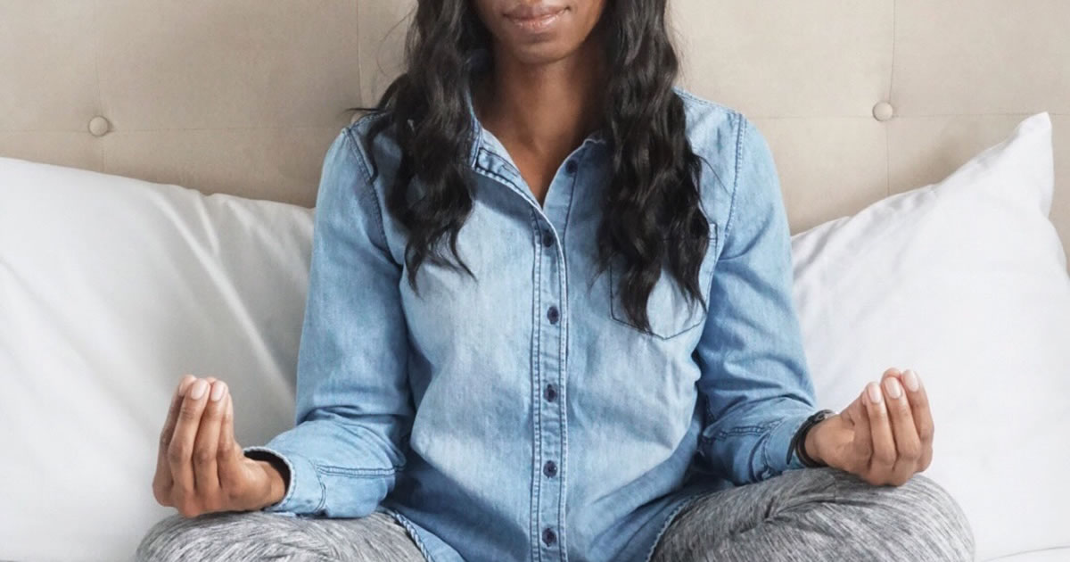 Woman sitting cross-legged on bed in meditative position with eyes closed and hands on knees