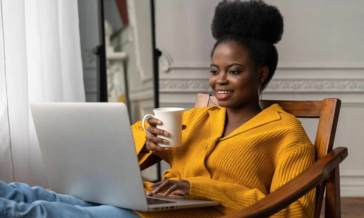 Mulher sorridente sentada em uma cadeira com as pernas para cima, usando um laptop e segurando uma caneca de café