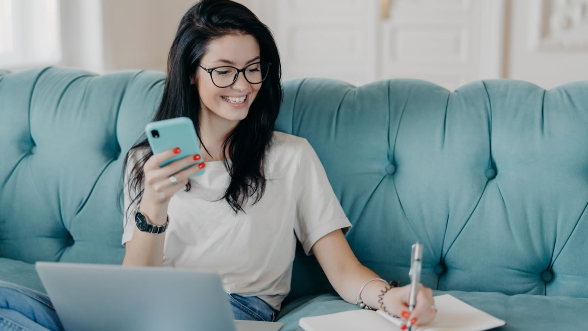 Smiling woman sitting on couch using mobile phone, laptop, pen, and notepad