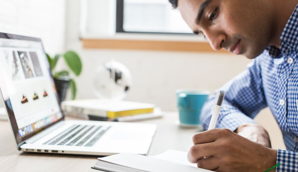 Man writing on notepad at desk with laptop