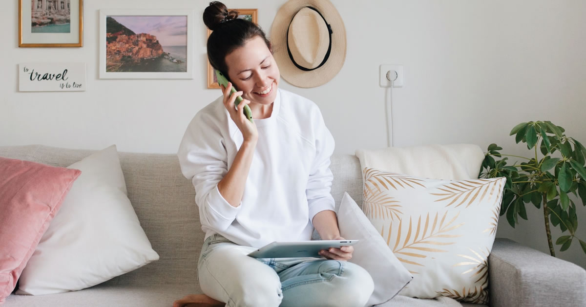 Young woman working from home, talking on a mobile phone and a tablet, smiling, sitting on the couch