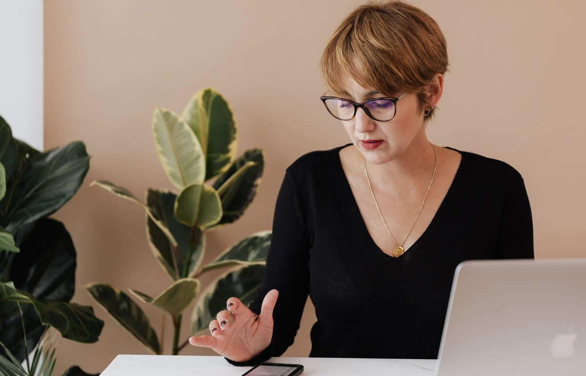 Woman writer studying mobile phone at desk with laptop