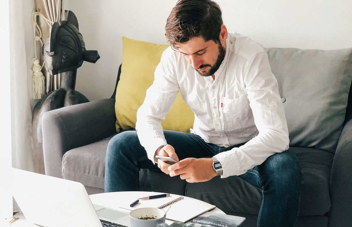 Young millennial working on his smartphone and laptop on a sofa