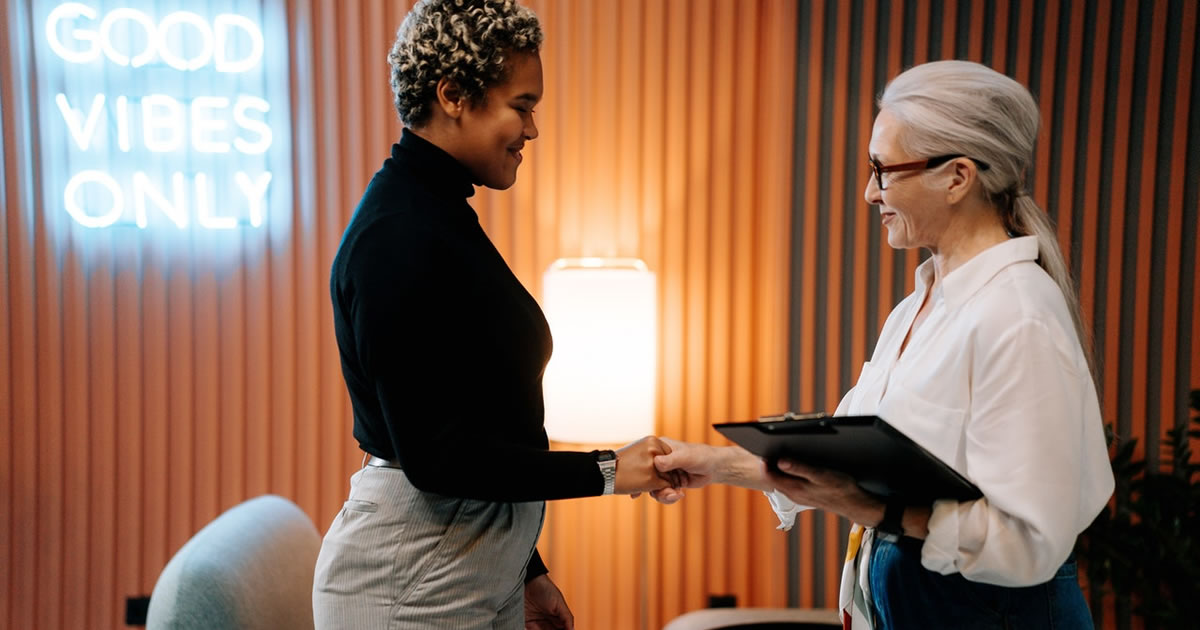 two women looking directly at each other, smiling and shaking hands