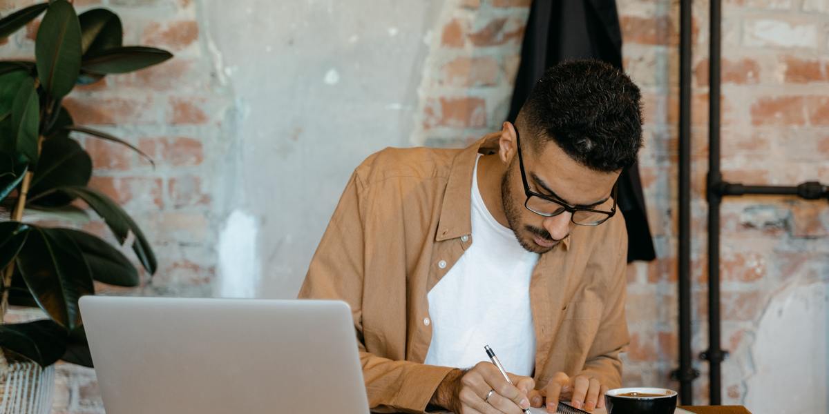 Homem em uma mesa com laptop, escrevendo em um caderno