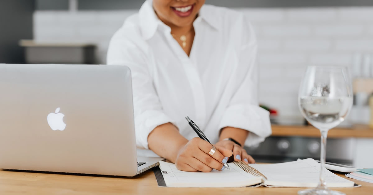 woman at table with laptop, writing in notebook