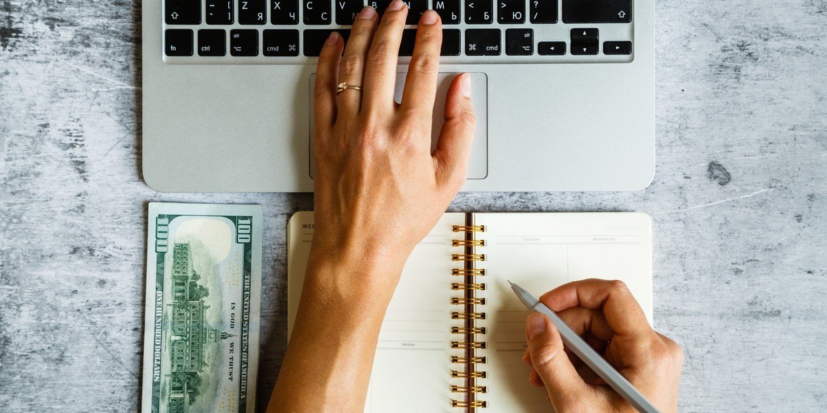 Writer holding pen and typing on laptop with notebook and one hundred-dollar bills lying on gray marble background