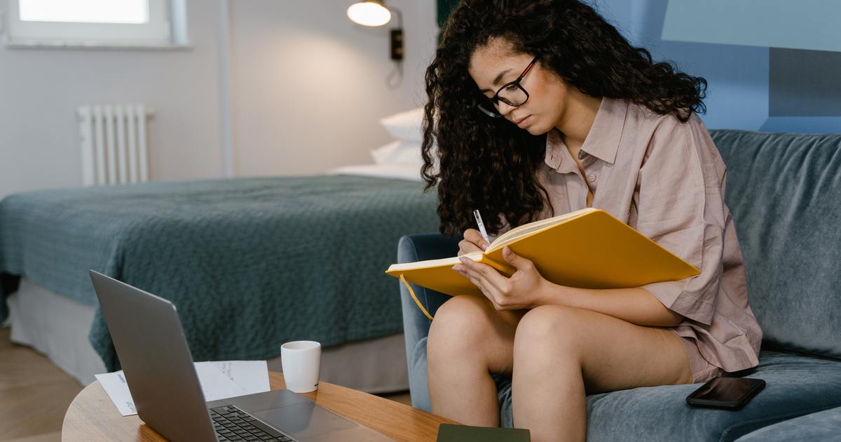 A woman sitting on a couch writing in a notebook near her laptop