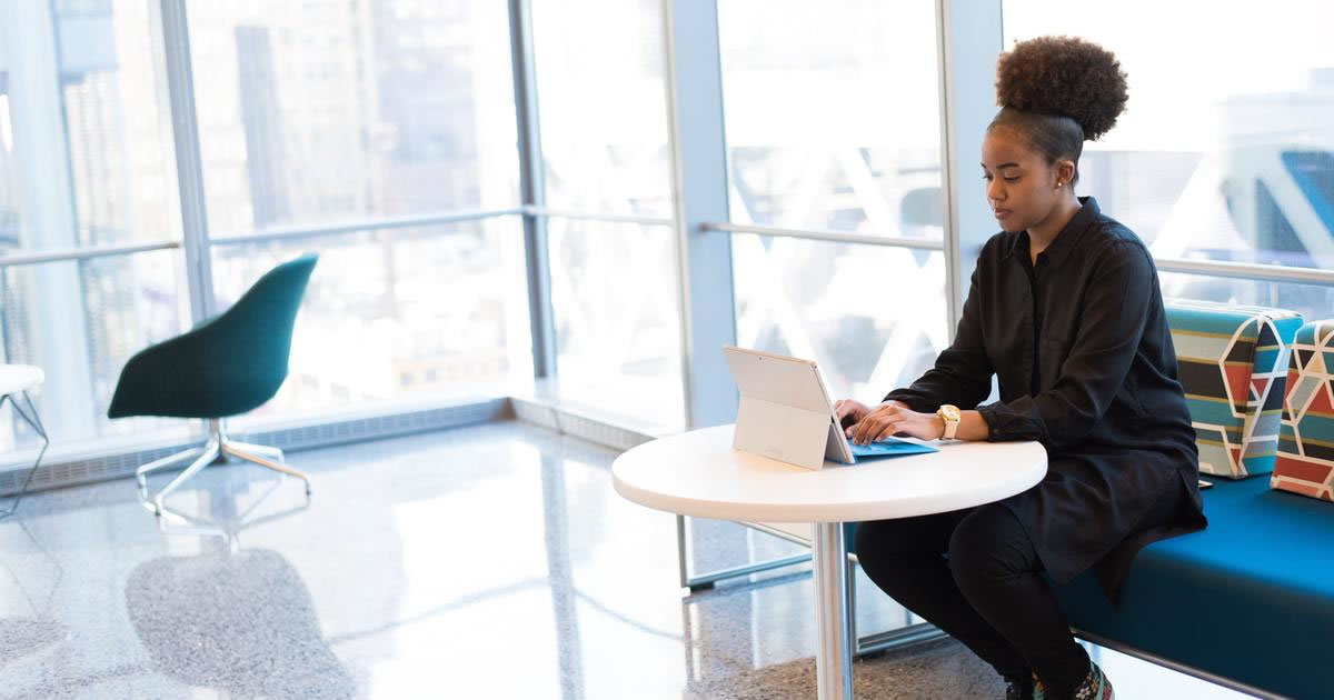 Woman seated at table in front of large windows typing on tablet