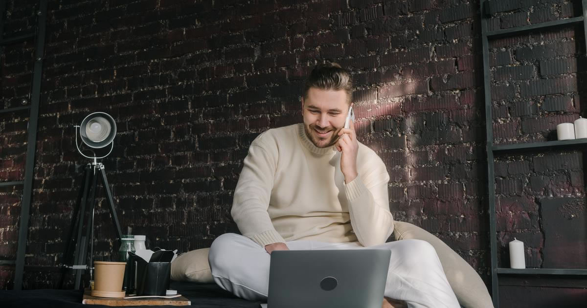 Smiling man seated at table talking on cell phone while looking at laptop screen