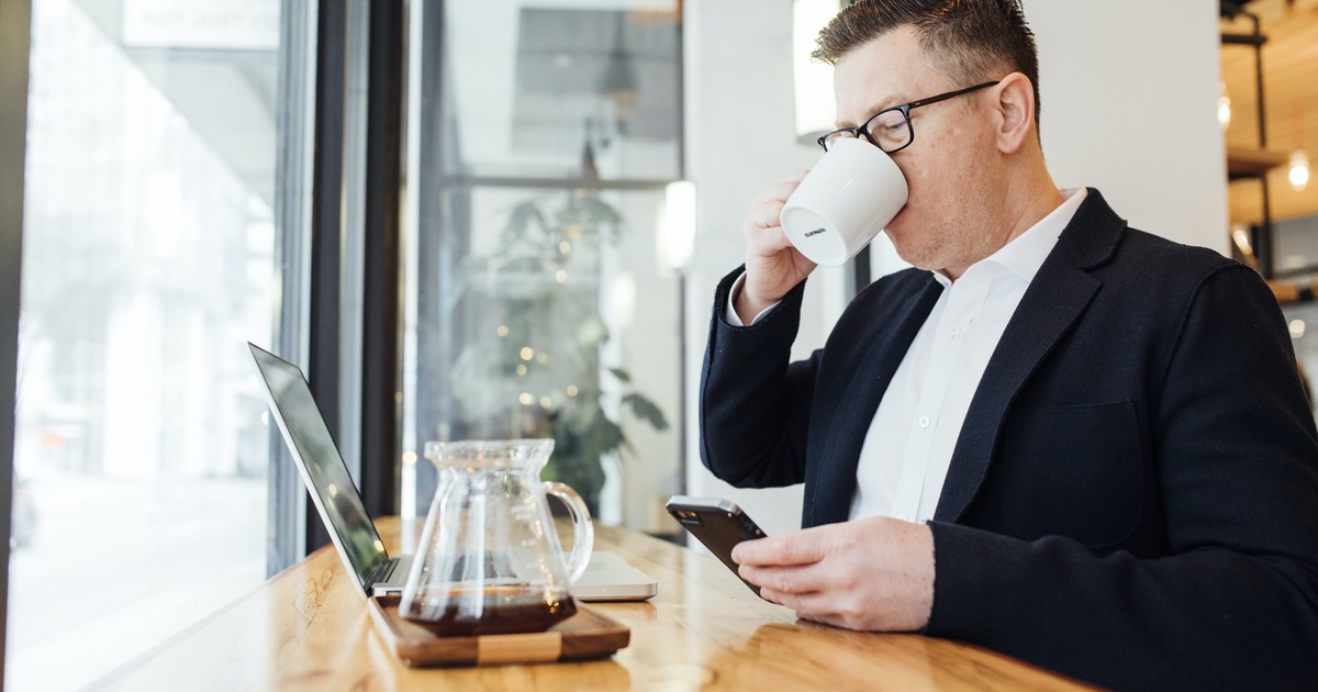 Empresário bebendo de uma caneca enquanto está sentado em frente ao laptop e ao celular